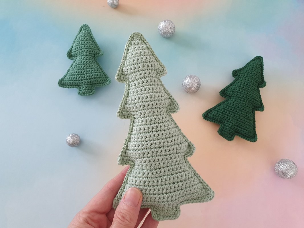 Nahaufnahme: Hand hält einen hellgrünen gehäkelten Weihnachtsbaum, zwei dunkelgrüne Bäume und silberne Kugeln auf pastellfarbenem Hintergrund