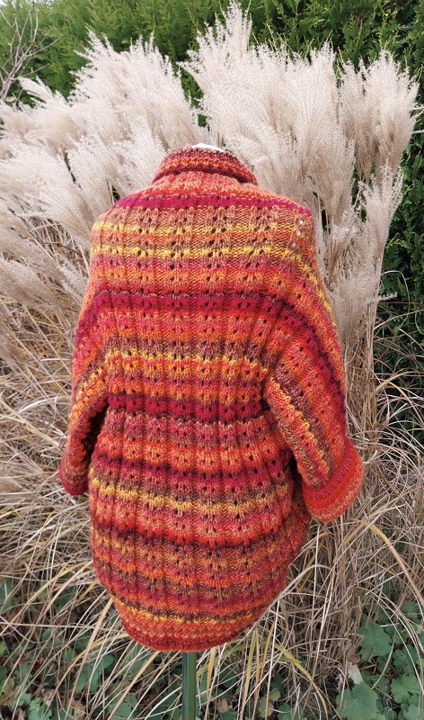 Rückansicht gestrickter Seelenwärmer in Rot‑Orange‑Gelb mit Streifen- und Lochmuster, auf Ständer vor Ziergras