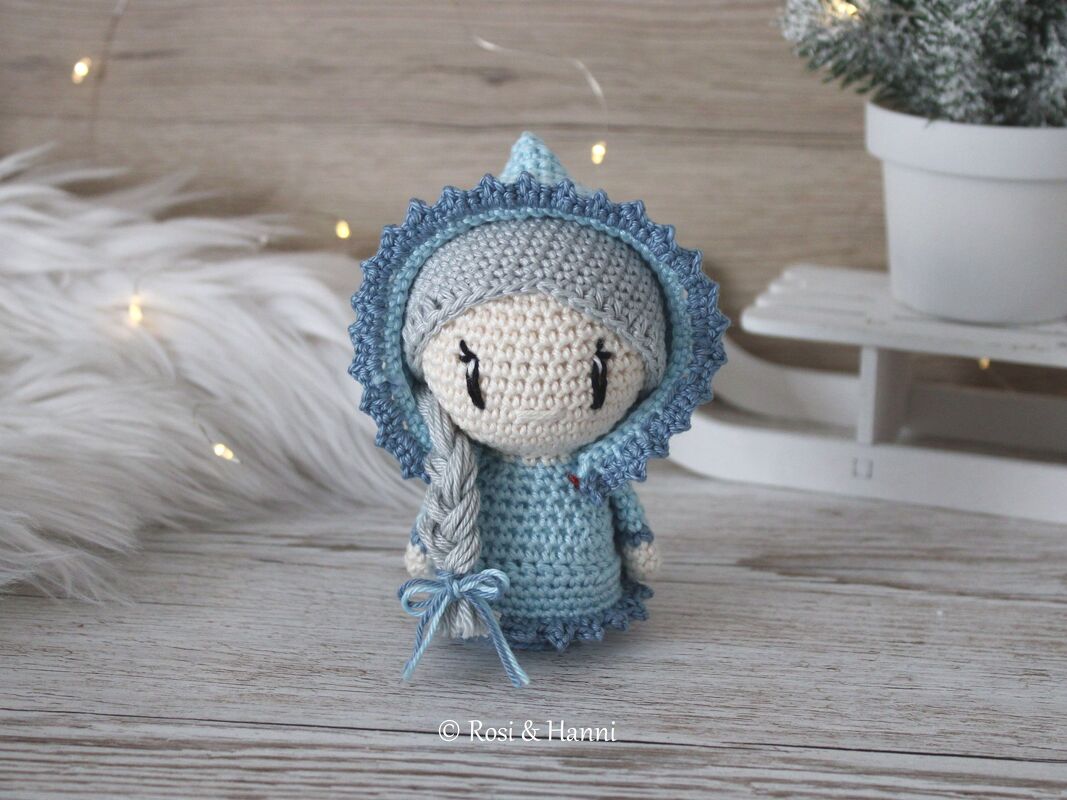 Gehäkelte Amigurumi‑Puppe in blauen und grauen Farbtönen mit Kapuze und geflochtenem Zopf auf hellem Holzuntergrund.
