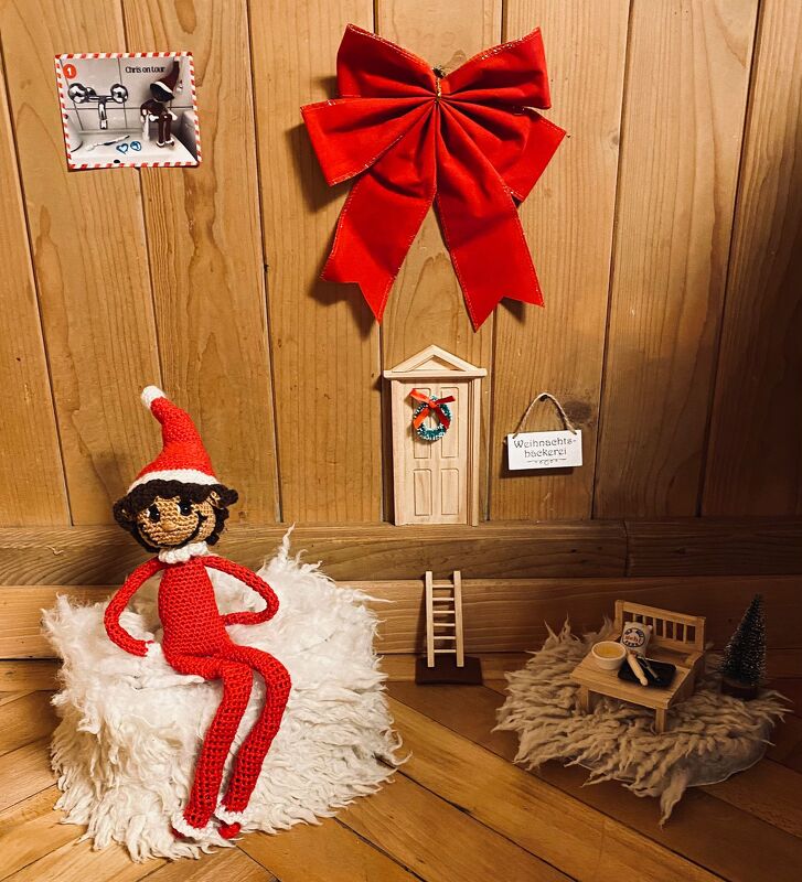 Gehäkelter Amigurumi-Wichtel in rotem Outfit auf flauschigem Hocker vor Holzwand mit roter Schleife und Miniaturtür