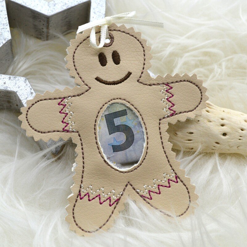 Beiger gestickter Lebkuchenmann-Anhänger mit ovalem Sichtfenster, darin ein gefalteter Geldschein, auf hellem, weichem Hintergrund