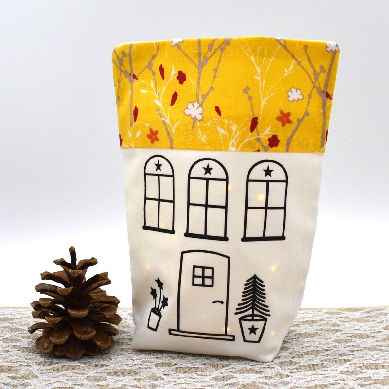Weißer Textilsack mit schwarzem Haus-Outline und gelbem, gemustertem Oberteil, innen von Lichtern erleuchtet, neben einem Tannenzapfen.
