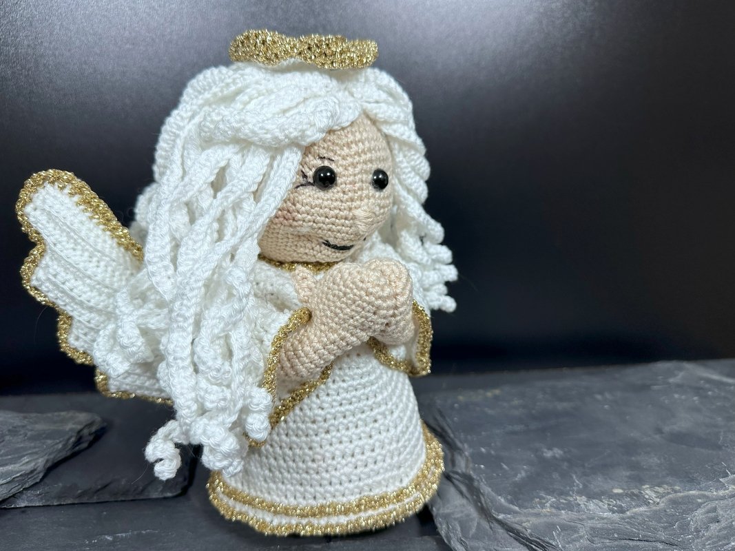 Gehäkelte Engelsfigur in Weiß mit goldenen Rändern, Heiligenschein und gefalteten Händen vor dunklem Hintergrund