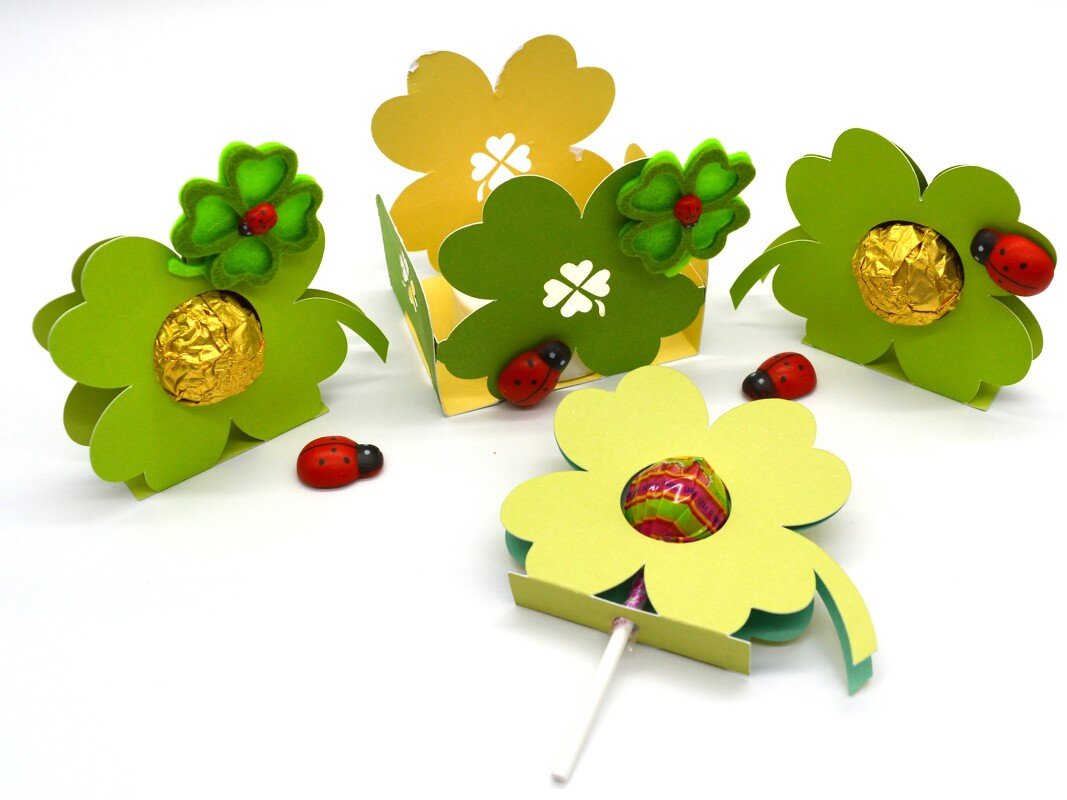 Mehrere vierblättrige Kleeblatt-Geschenkboxen und ein Lutscherhalter aus grünem und gelbem Papier, dekoriert mit Filzkleeblättern und kleinen Marienkäfern; in einigen Boxen sind goldene Pralinen zu sehen.