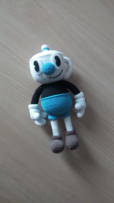A crocheted amigurumi doll with a white cup-like head, blue nose, black torso, blue shorts, white hands, and brown shoes on a light wooden surface.