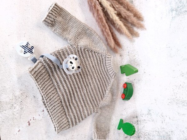 Beiger gestrickter Baby-Pullover mit Rippenmuster, Schnullerhalter und Holzspielzeug auf hellem Untergrund