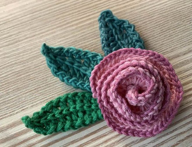 Gehäkelte rosa Rose mit drei grünen Blättern auf Holzoberfläche.