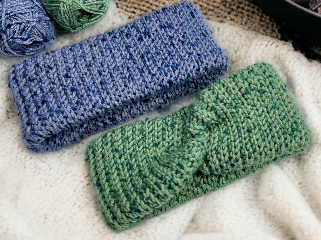 Two speckled knit-look headbands, one blue and one green with a twisted center, resting on a cream blanket with yarn balls nearby.