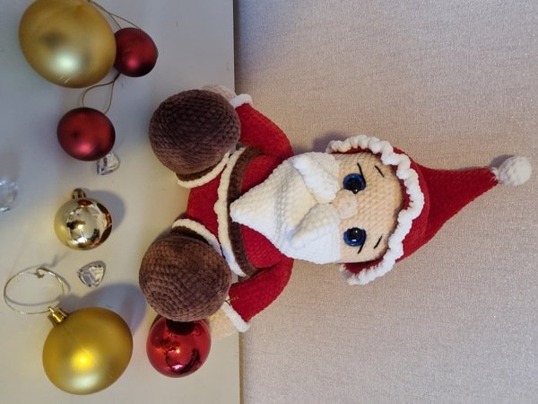Gehäkelter Weihnachtsmann mit roter Mütze, weißem Bart und braunen Handschuhen neben goldenen und roten Kugeln