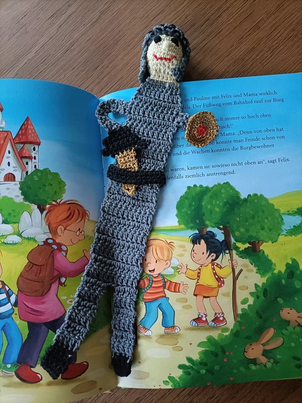 Handgehäkeltes Ritter-Lesezeichen in Grau mit Gesicht, Helm und rundem Schild auf geöffneter Kinderbuchseite