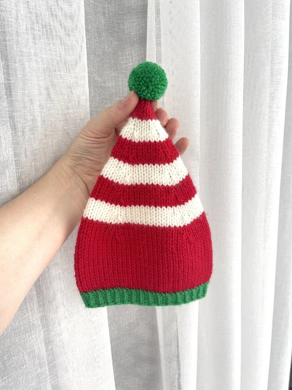 Pointed red and white striped knit beanie with green ribbed cuff and green pom-pom held by hand