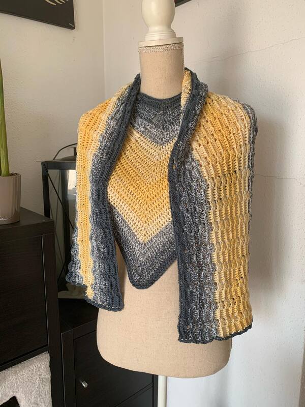 Gehäkelter Wickelschal in Gelb und Grau mit V-förmigem Chevron-Mittelteil und texturierten Seiten auf einer Schneiderpuppe.