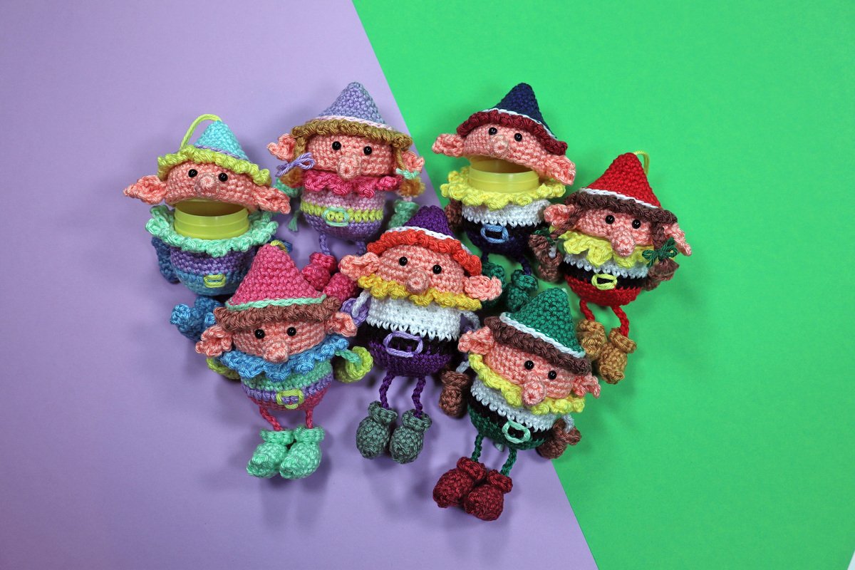 Mehrere bunte gehäkelte Amigurumi‑Wichtel mit Aufhängeschlaufen auf zweifarbigem Hintergrund