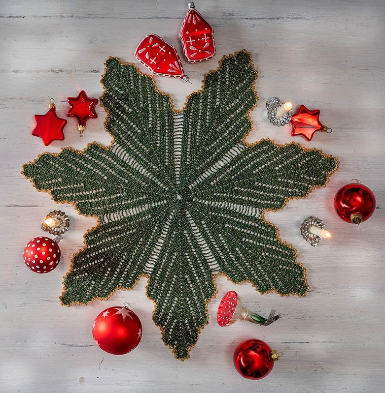 Sternförmiges grünes Häkeldeckchen mit goldener Kante auf hellem Holz, umgeben von roten Christbaumkugeln und kleinen Kerzen.