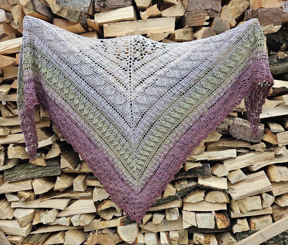 Crocheted triangle shawl with gradient from light grey to green to purple and varied stitch patterns, draped over stacked firewood.