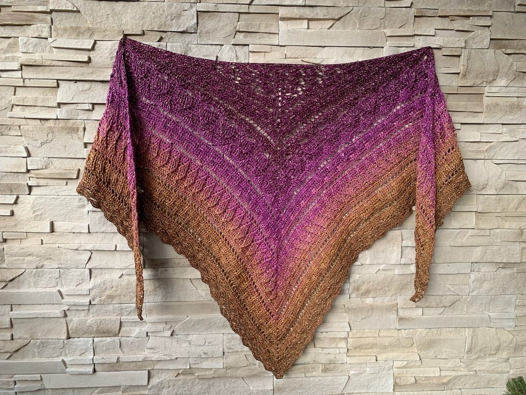 Triangle crochet shawl in purple fading to bronze, hung against a neutral stone wall