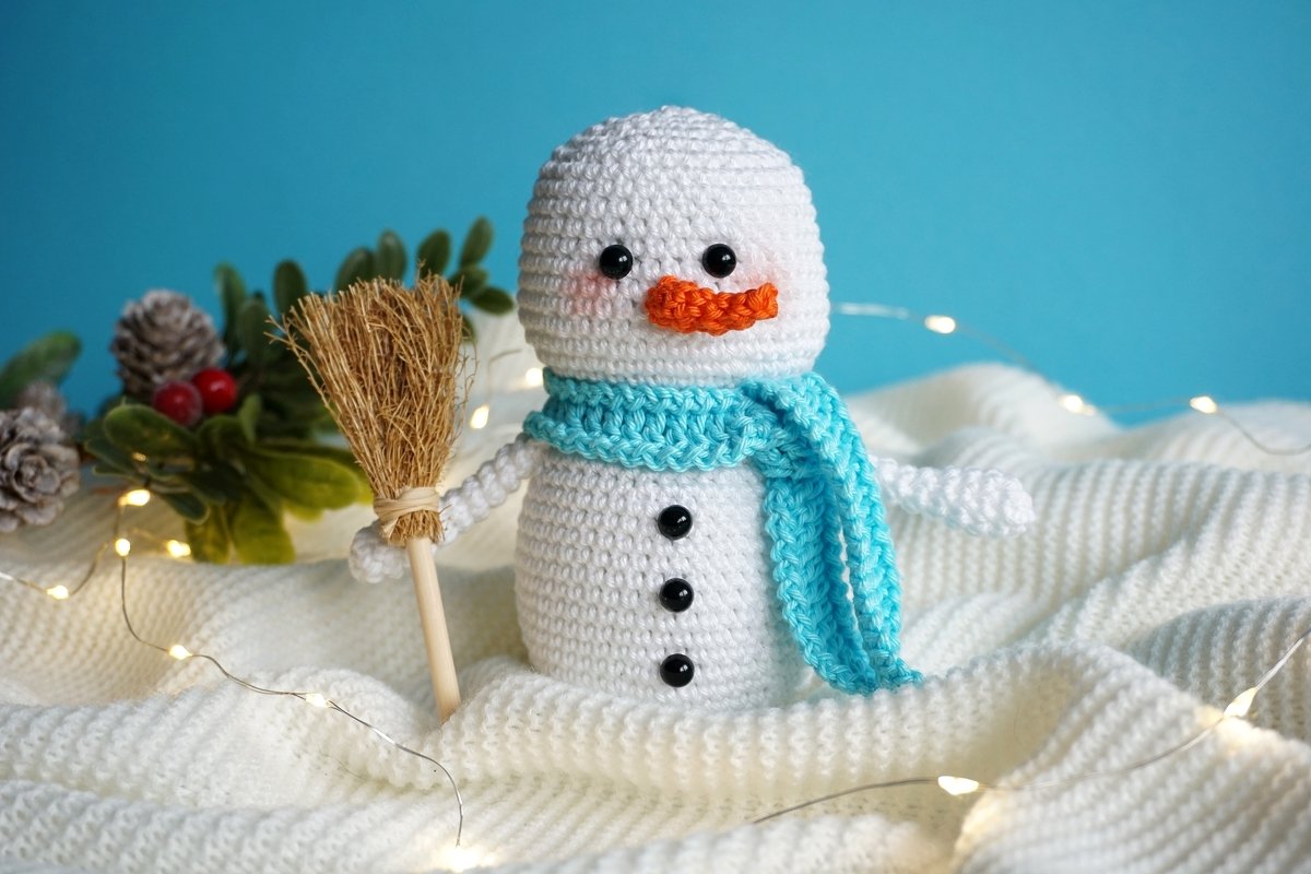 Gehäkelter weißer Amigurumi-Schneemann mit blauem Schal und Besen auf weißer Decke, Lichterkette und Tannengrün im Hintergrund