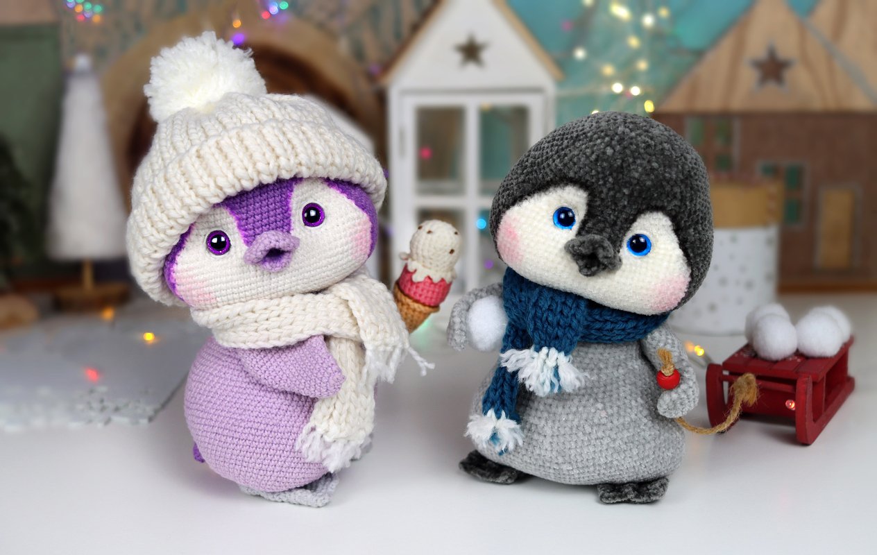 Two crocheted penguin amigurumi — lavender with cream hat and scarf holding an ice-cream prop, and gray with blue scarf beside a small red sled with snowballs