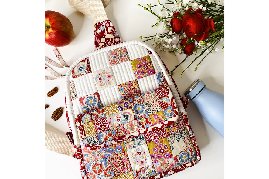 Nahaufnahme eines gesteppten Mini-Rucksacks mit buntem Blumen-Patchwork und vorderem Fach, daneben Apfel, Nüsse und Blumen.
