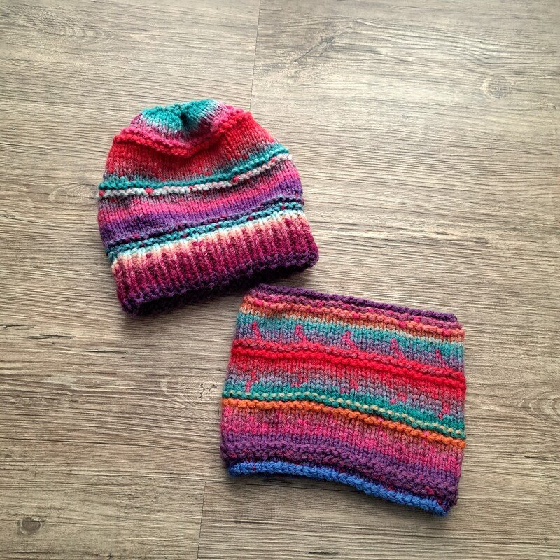 Gestrickte Kinder‑Mütze und passender Loop in buntem Farbverlauf auf Holzboden