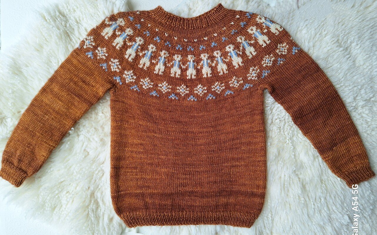 Gestrickter rostbrauner Kinderpullover mit mehrfarbiger Rundpasse aus weißen und hellblauen Figuren- und Schneeflockenmotiven, liegend auf hellem Plüschuntergrund.