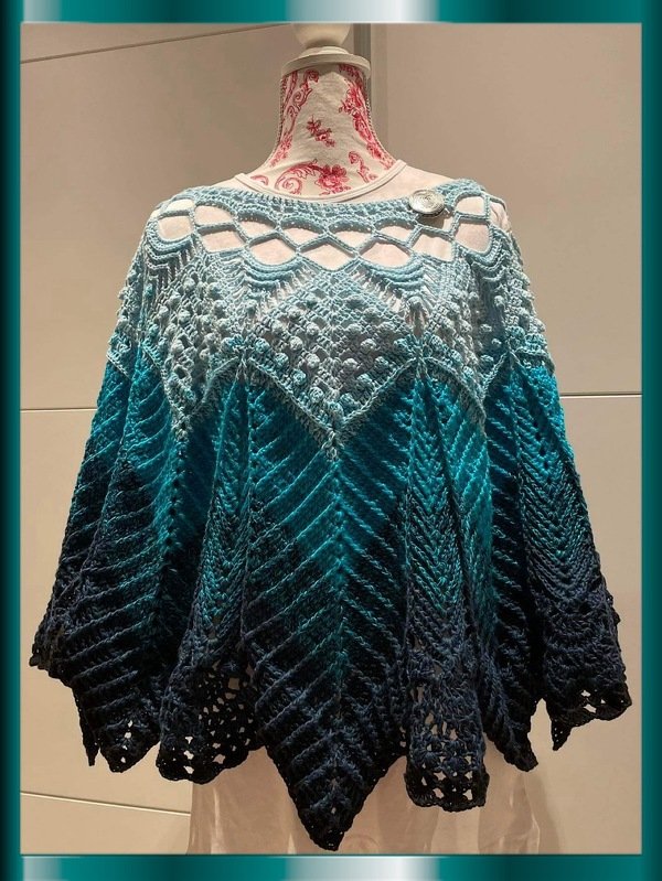 Gehäkelter Poncho in abgestuften Blautönen mit oberem Netzmuster, texturierten Maschen und gezacktem Saum auf einer Schaufensterpuppe.