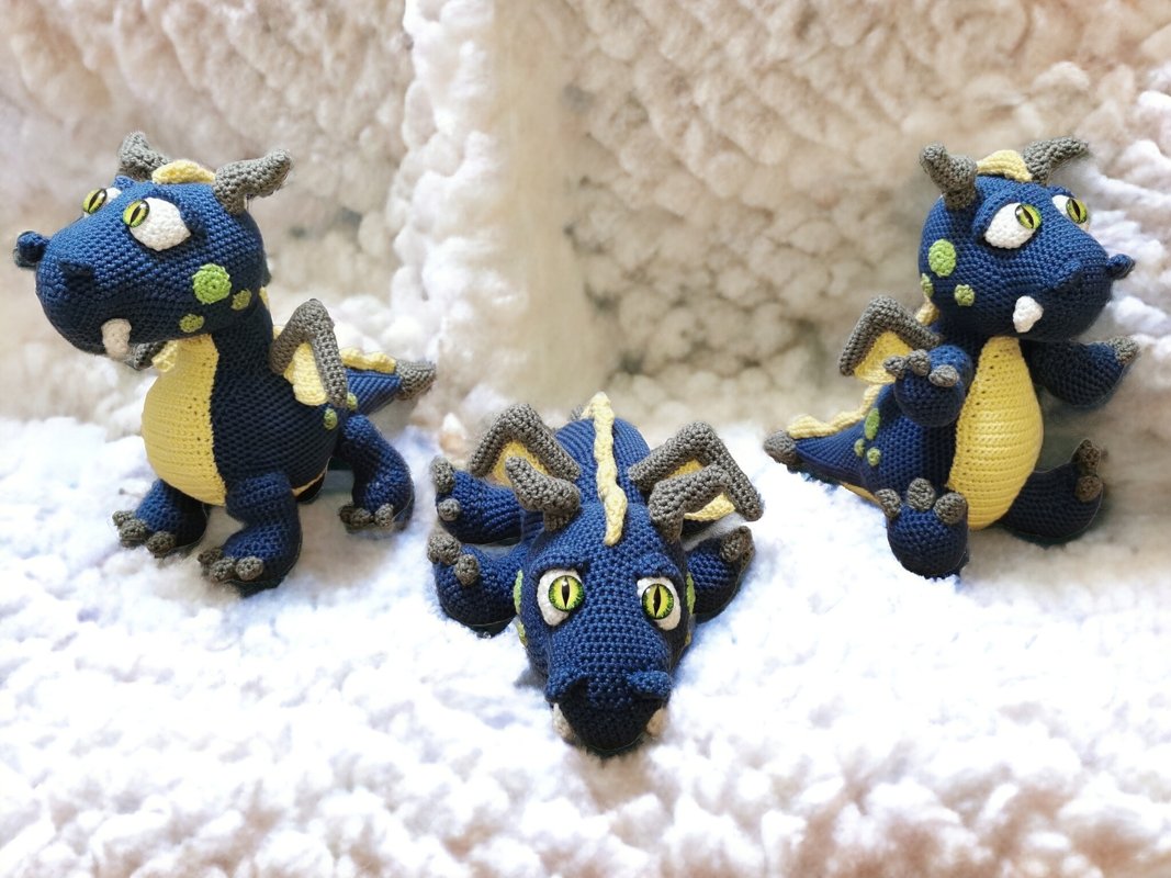 Three crocheted blue dragons with yellow bellies and grey horns shown from front, side and top head views on a soft white background