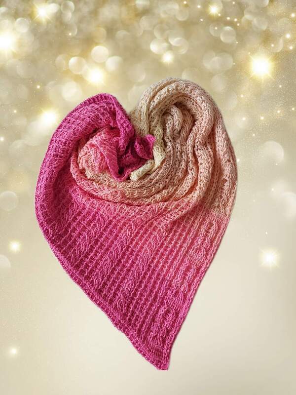 Gehäkeltes Dreieckstuch in Pink- und Beigetönen, zu einem Herz gelegt vor goldenem Bokeh-Hintergrund.