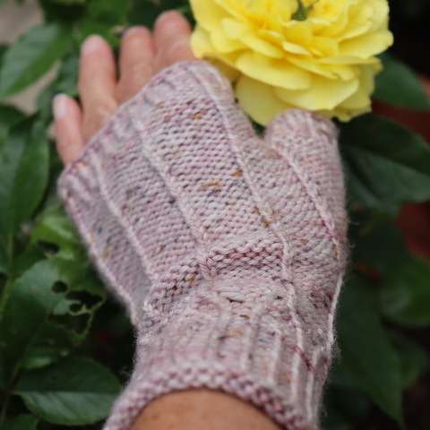Hand mit pastellrosa gestrickter, fingerloser Handstulpe und gelber Rose im Hintergrund