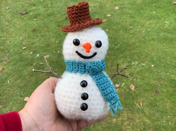Hand hält handgehäkelten weißen Schneemann mit braunem Hut und türkisfarbenem Schal vor grünem Rasen