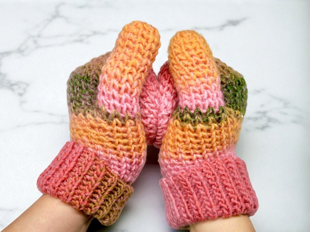 Hände in gehäkelten Fäustlingen mit weichem Farbverlauf aus Rosa, Orange und Grün.