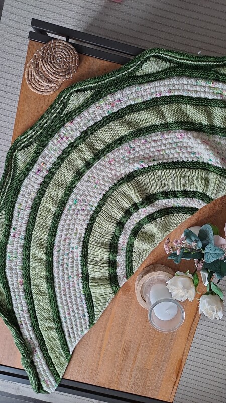 Halbrundes gestricktes Tuch in grünen und cremefarbenen Streifen auf einem Holztisch neben Kerzen und Blumen