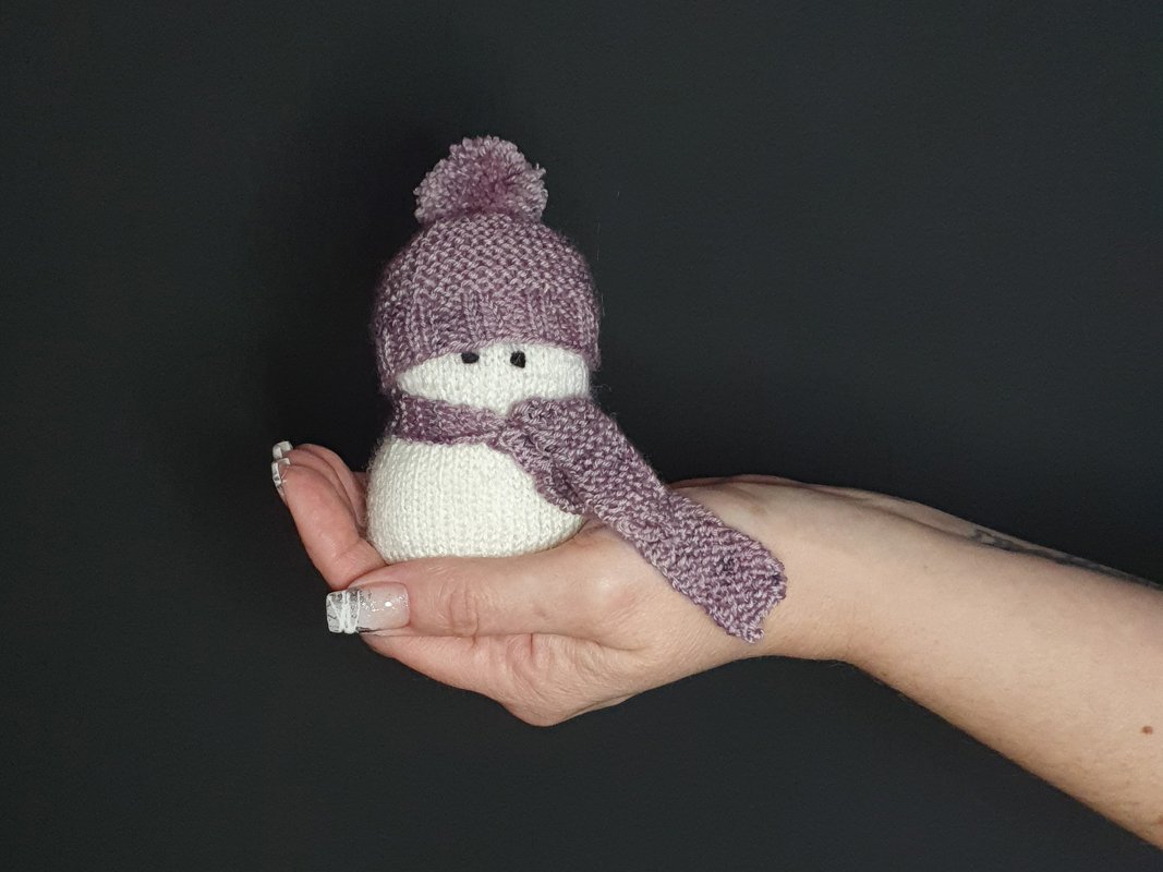 Hand hält kleinen gestrickten Schneemann mit fliederfarbener Mütze und Schal