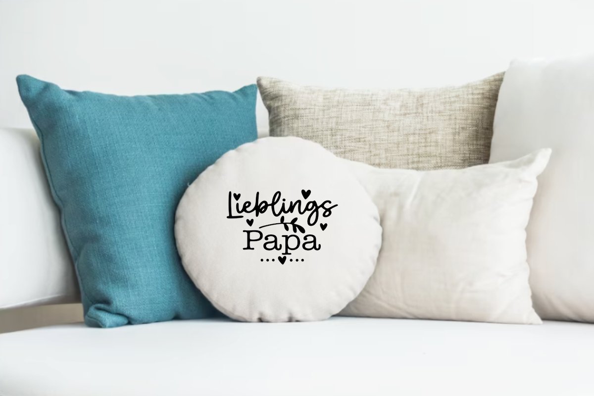 Rundes cremefarbenes Kissen mit schwarzem Schriftzug „Lieblings Papa“ und Herzmotiven auf einem Sofa.
