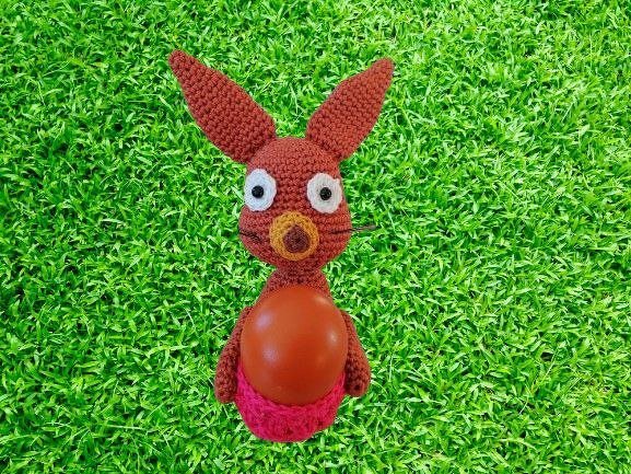 Brauner gehäkelter Amigurumi-Hase mit langen Ohren, weißen Augen und orangefarbenem Ei auf grünem Gras