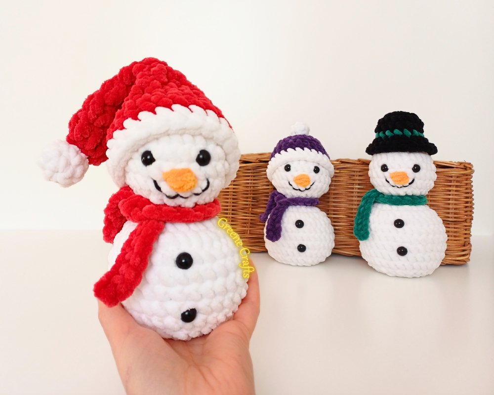 Three crocheted amigurumi snowmen with varied hats and scarves on a wicker tray, one positioned in front and held slightly by a hand.