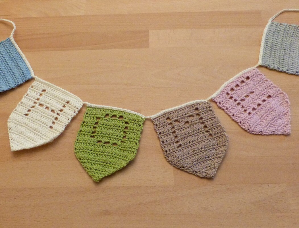 Gehäkelte Wimpelkette mit pastellfarbenen Wimpeln auf Holzuntergrund