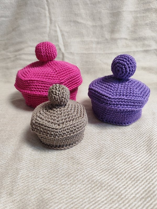 Drei kleine gehäkelte Dosen mit Deckeln und kugelrunden Griffen in Fuchsia, Lila und Taupe auf hellem Stoff