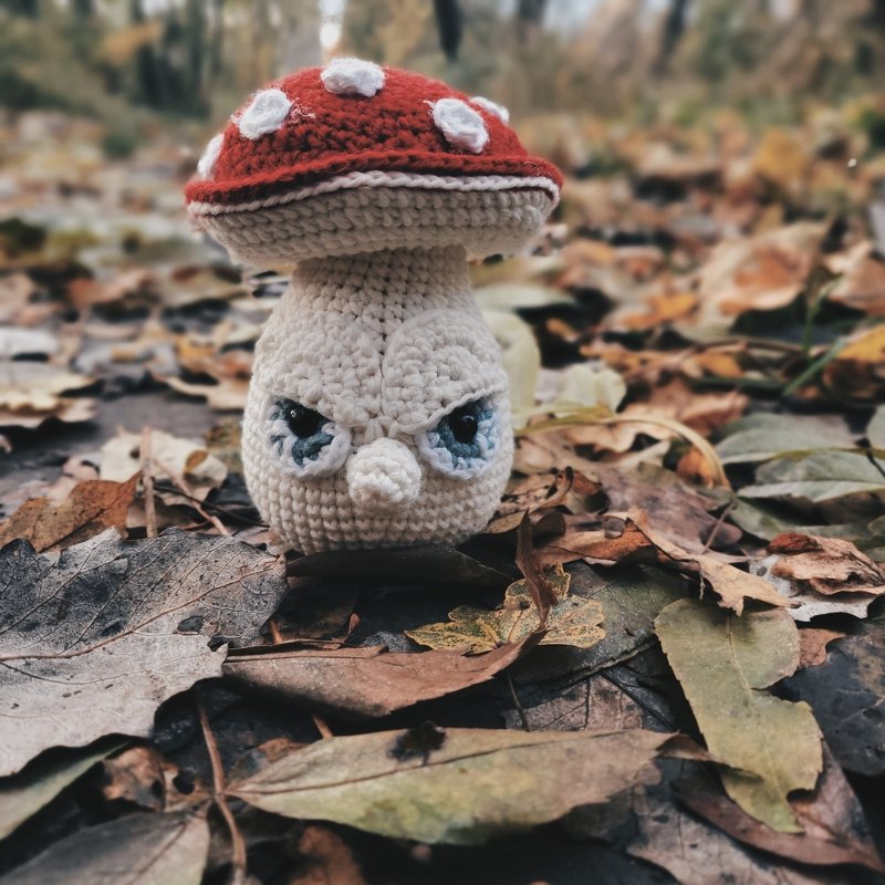 Gehäkelter Amigurumi-Pilz mit rotem Hut und mürrischem Gesicht auf braunem Herbstlaub