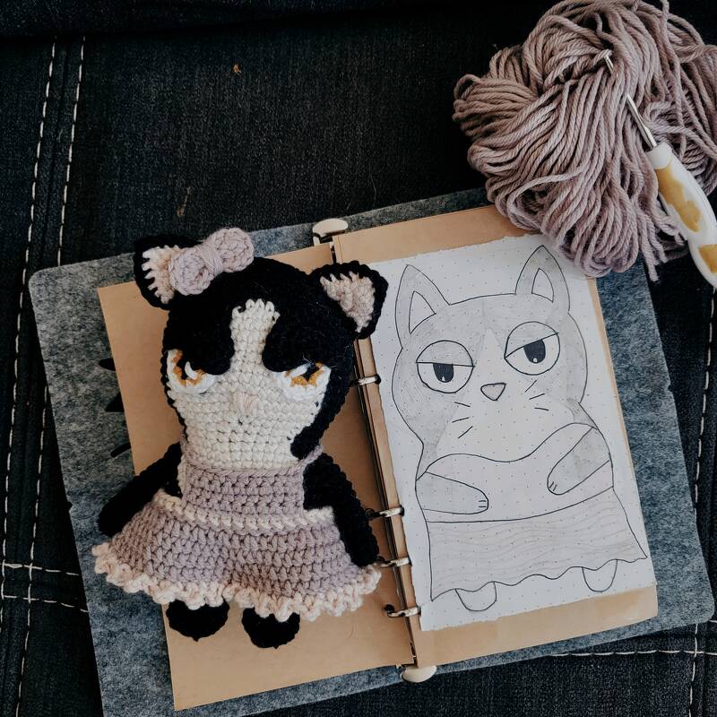 Gehäkelte Amigurumi-Katze neben Notizbuch-Skizze, Garnknäuel und Häkelnadel auf Stoffuntergrund