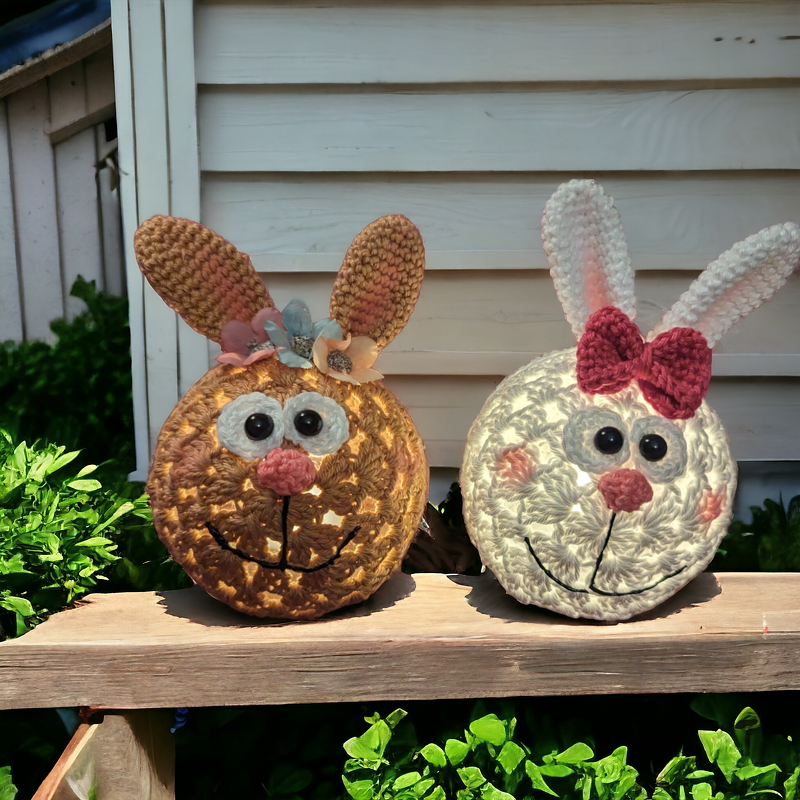 Zwei gehäkelte Hasenköpfe als Lichter, braun mit Blumen und weiß mit rosa Schleife, auf einer Holzleiste.