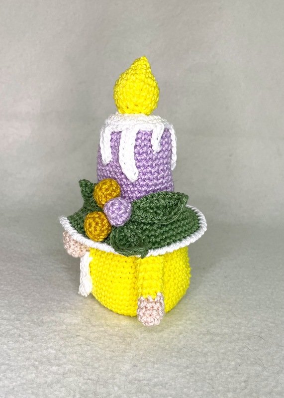Gehäkelter Amigurumi-Wichtel mit gelbem Körper, lila Kerzenhut, weißem Wachs, grünen Blättern und gelber Flamme