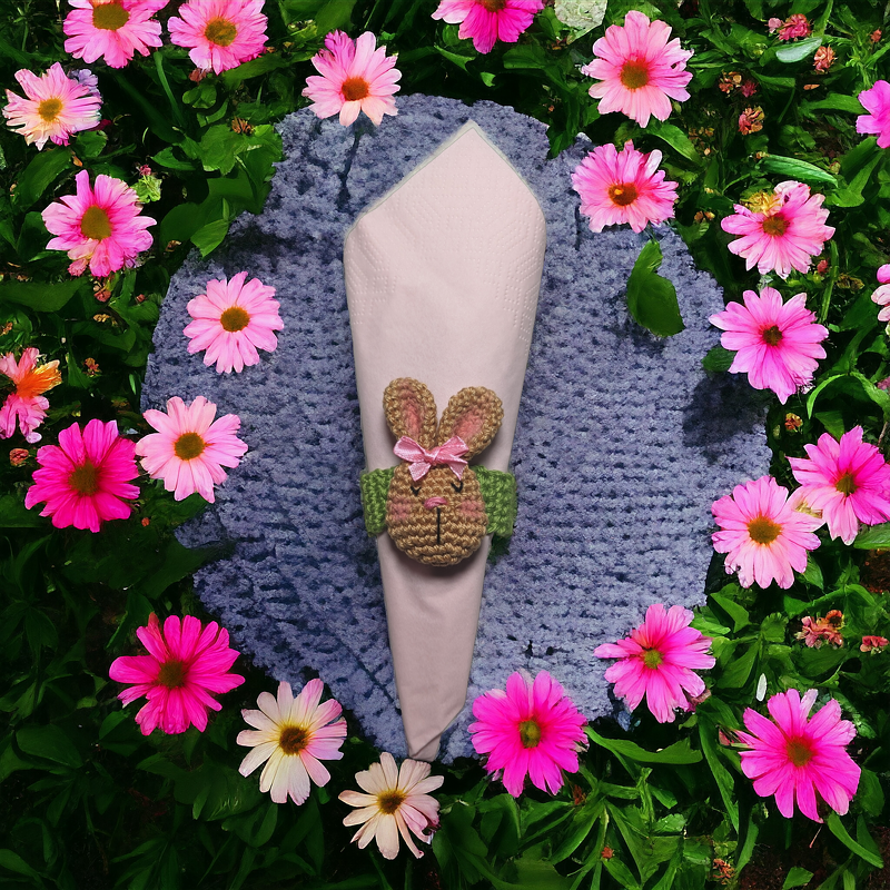 Gehäkelter Hasen-Serviettenring um eine rosa Serviette auf blauem Häkeldeckchen, umgeben von rosa Blumen