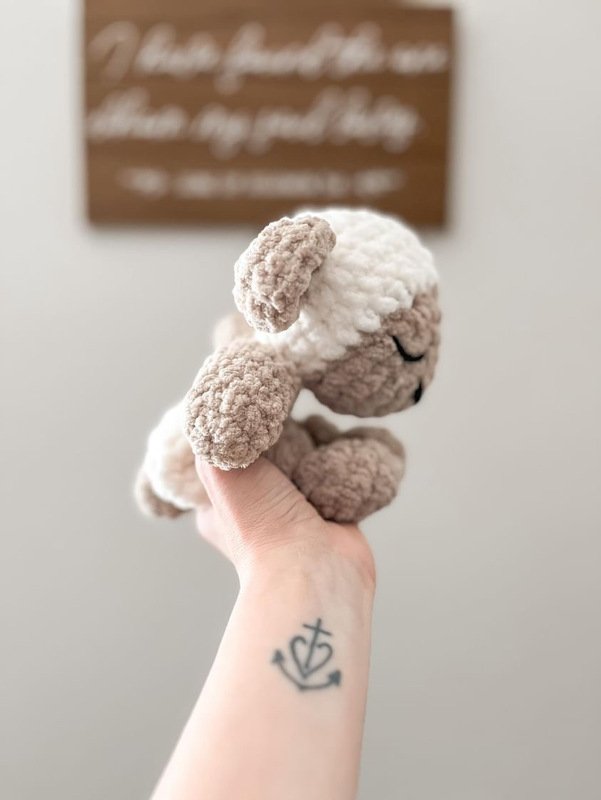 Hand holding a small cream-and-beige crocheted lamb; an anchor tattoo is visible on the wrist.