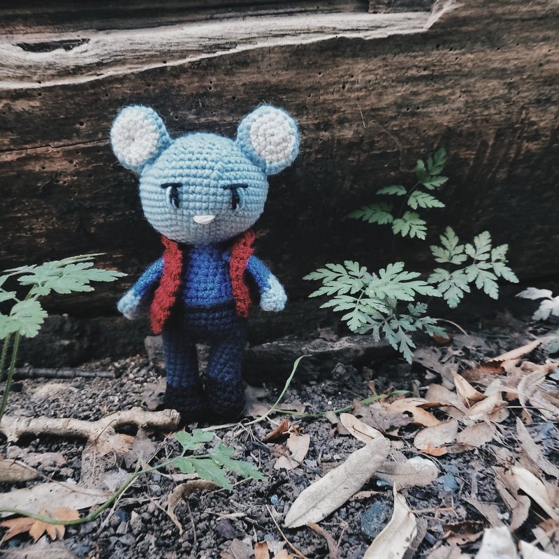 Gehäkelte hellblaue Amigurumi‑Maus mit roter Weste vor Baumstamm