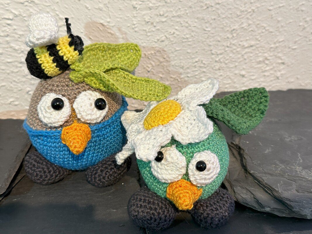 Zwei gehäkelte Amigurumi-Eulen, braun mit Biene, grün mit Gänseblümchen