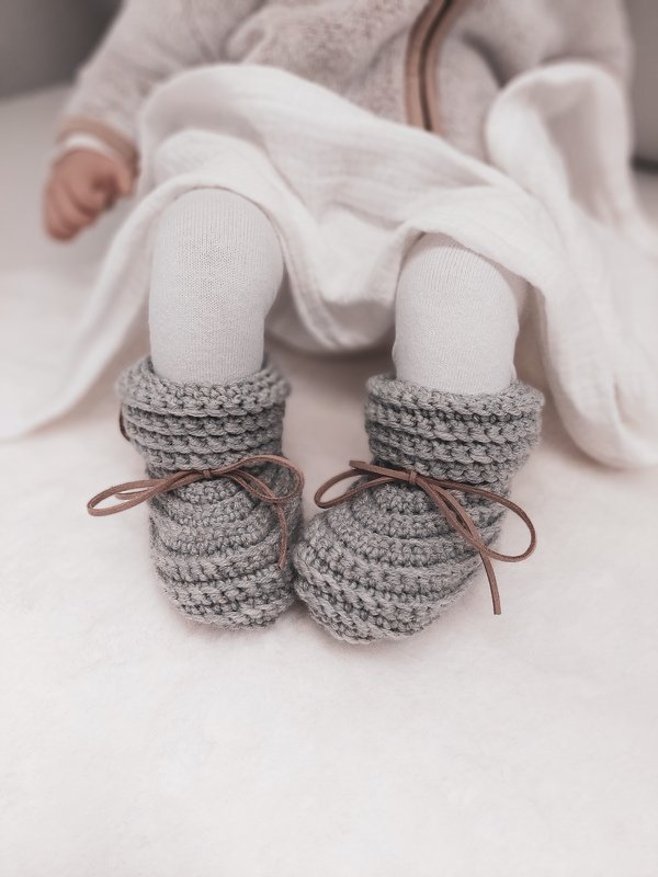 Gehäkelte graue Babyschuhe mit braunen Schnürbändern an Babyfüßen auf hellem Stoff