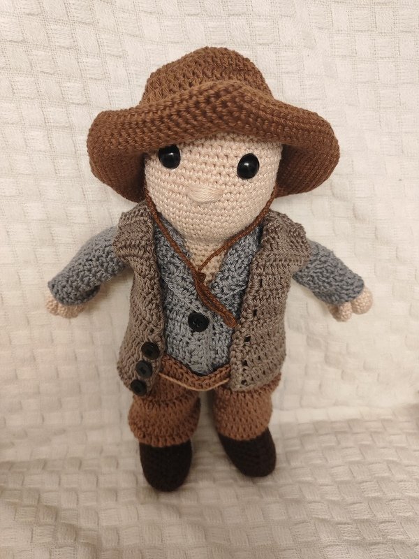 Gehäkelte Amigurumi-Puppe mit braunem Hut, grauem Oberteil, brauner Weste, Hose und dunklen Stiefeln vor hellem Stoffhintergrund
