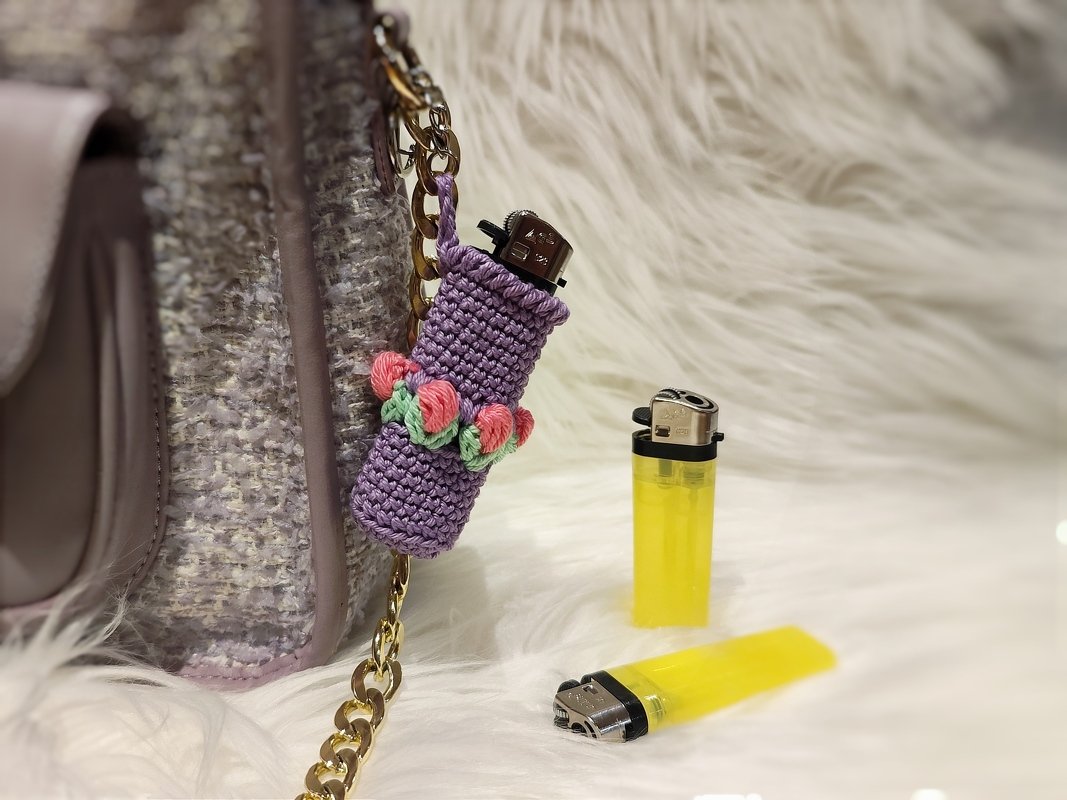 Lila gehäkelter Feuerzeughalter mit rosa-grünen Blüten an goldfarbener Kette, daneben zwei gelbe Feuerzeuge auf weißem Kunstfell.