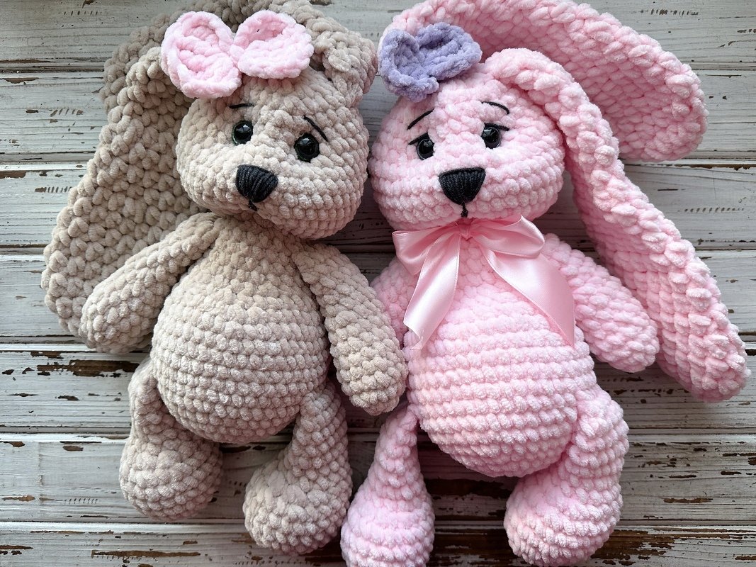 Two crocheted amigurumi bunnies in beige and pink with bows and a satin neck ribbon.